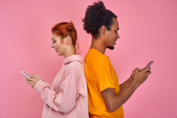 Beautiful, pretty, cute red hair woman and handsome dark skinned man standing back to back and writing messages through 4g internet on modern smartphones, enjoy nice conversation over pink background.