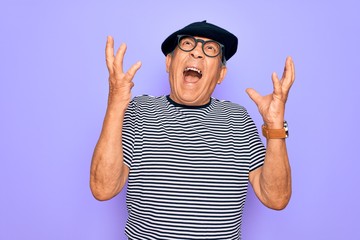 Senior handsome hoary man wearing french beret and glasses standing over purple background crazy...