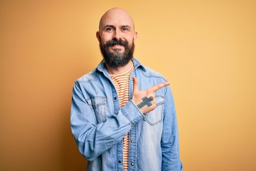 Handsome bald man with beard wearing casual denim jacket and striped t-shirt cheerful with a smile of face pointing with hand and finger up to the side with happy and natural expression on face