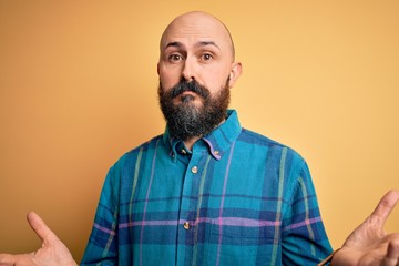 Obraz premium Handsome bald man with beard wearing casual shirt standing over isolated yellow background clueless and confused expression with arms and hands raised. Doubt concept.