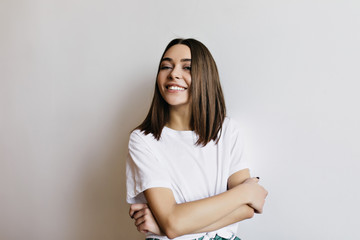 Obraz premium Pleasant girl with shiny brown hair smiling on white background. Indoor photo of caucasian lady standing in studio with arms folded.