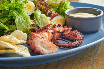 Appetizing salad with grilled octopus tentacle, quail eggs, lettuce, croutons and white sauce in a blue plate on a wooden background. Close up selective focus