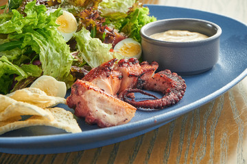 Appetizing salad with grilled octopus tentacle, quail eggs, lettuce, croutons and white sauce in a blue plate on a wooden background. Close up selective focus