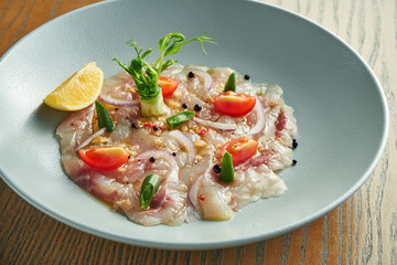 Chilled sea bass carpaccio with red onion, cherry tomatoes and hot chili peppers in a beige bowl on a wooden background. Close up, selective focus