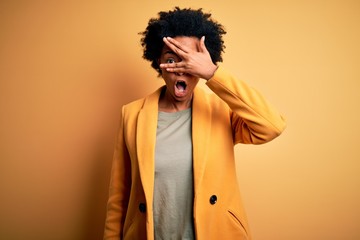 Fototapeta na wymiar Young beautiful African American afro businesswoman with curly hair wearing yellow jacket peeking in shock covering face and eyes with hand, looking through fingers with embarrassed expression.