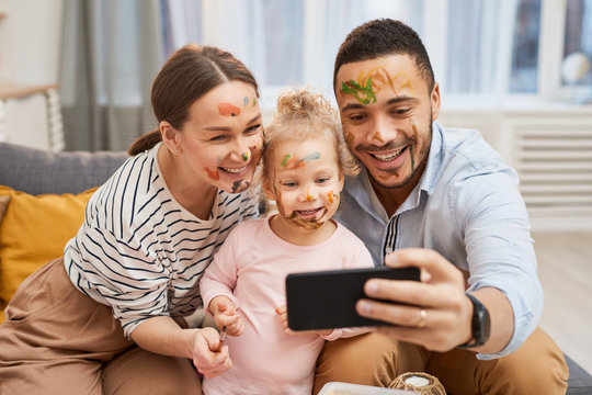 Young man and woman taking funny selfie on smartphone with their little daughter with gouache on their faces
