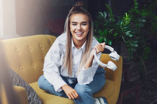 Woman sits on yellow sofa and holds in her hand white wireless headphones. Millennial girl looks the camera with smile