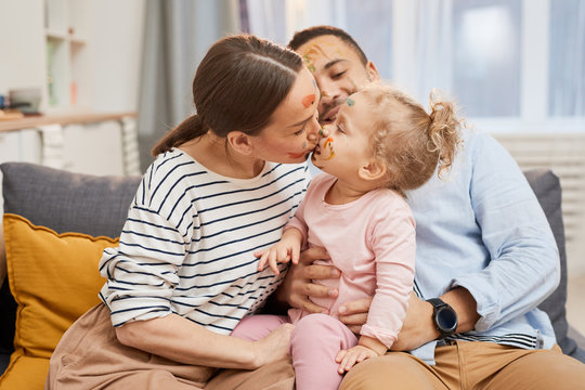 Happy young family of three sitting together on sofa, mother kissing her little daughter, horizontal medium shot