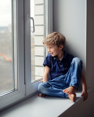 Little cute boy sits on a window sill, looks out the window and smiles.