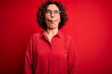 Obraz na płótnie Canvas Middle age beautiful curly hair woman wearing casual shirt and glasses over red background making fish face with lips, crazy and comical gesture. Funny expression.