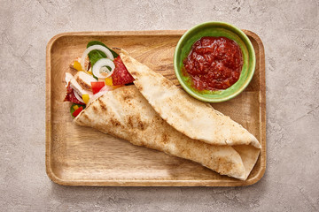 top view of fresh burrito with chicken and vegetables on board near chili sauce on concrete grey background