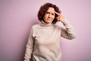 Fototapeta na wymiar Middle age beautiful curly hair woman wearing casual turtleneck sweater over pink background pointing unhappy to pimple on forehead, ugly infection of blackhead. Acne and skin problem