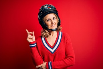 Middle age beautiful blonde motorcyclist woman wearing moto helmet over red background with a big smile on face, pointing with hand and finger to the side looking at the camera.
