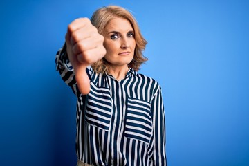 Middle age beautiful blonde woman wearing casual striped shirt standing over blue background looking unhappy and angry showing rejection and negative with thumbs down gesture. Bad expression.