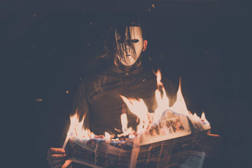 A man in black shirt and white mask reading a burning newspaper. Black hair partly covers the white...