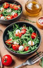Healthy arugula salad with mozzarella cheese and cherry tomatoes in black bowls on beige rustic background.