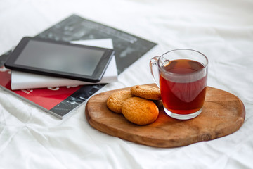 A Cup of tea, cookies on a wooden surface. A tablet computer, a magazine, and a book on the bed. Morning news. 