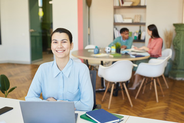 Cheerful young adult businesswoman sitting at office desk with laptop on it smiling at camera, her colleagues working behind her