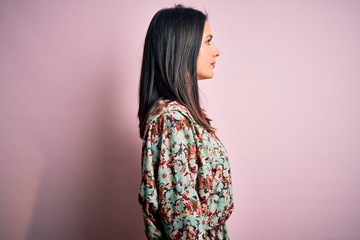 Young brunette woman with blue eyes wearing floral colorful dress over pink background looking to side, relax profile pose with natural face with confident smile.