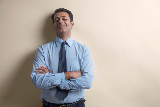 Smiling businessman with eyes closed standing with his back against a wall with arms crossed
