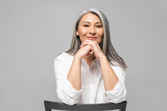 beautiful positive asian woman with grey hair sitting on chair isolated on grey