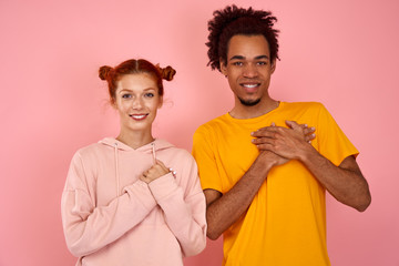 Kind hearted friendly looking young red haired woman and black man keep both palms on chest, dressed casually, express gratitude and good feeling, stand next to each other, over pink studio background