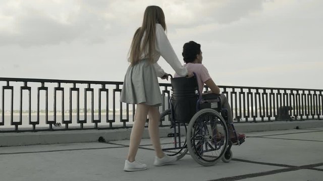 Young disabled man is walking using wheelchair, woman is rolling it. They are resting together in park near river