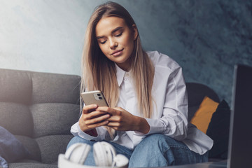 Obraz premium Pretty casually dressed girl with long hair sitting on coach and texting on smartphone. Young lady using portable mobile