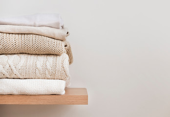 A stack of neatly folded warm knitwear, wool on a wooden shelf and a beige background. Capsule...