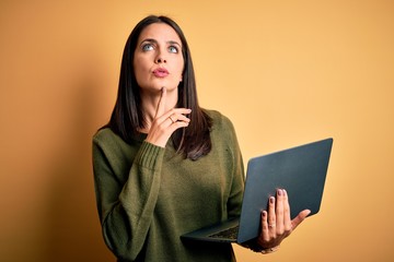 Young brunette woman with blue eyes working using computer laptop over yellow background Thinking concentrated about doubt with finger on chin and looking up wondering