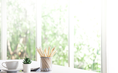 Office desk decorate with a cup of coffee, small beautiful green tree, calculator and pencils in metal basket. Morning sunlight. Free copy space on right for design or text.