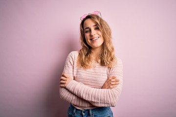 Young beautiful blonde woman wearing casual sweater and sunglasses over pink background happy face smiling with crossed arms looking at the camera. Positive person.