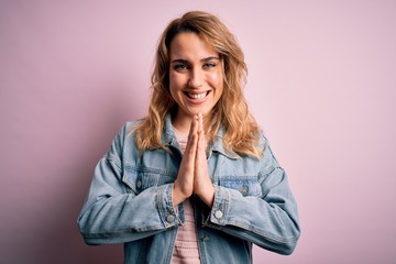 Young beautiful blonde woman wearing casual denim jacket standing over pink background praying with hands together asking for forgiveness smiling confident.