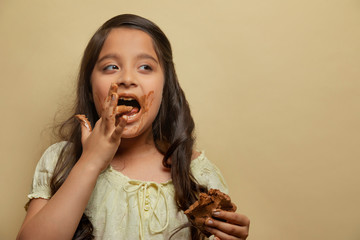 Girl licking chocolate from fingers holding a chocolate bar in the other hand looking away
