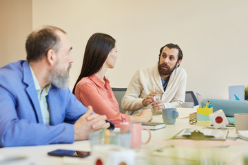 Young bearded man sitting at office table in board room with his colleagues sharing his ideas, copy space