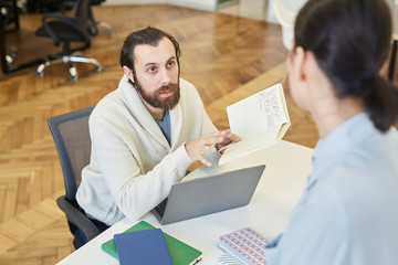Young man sitting at office desk sharing his ideas to unrecognizable colleague, horizontal high angle shot
