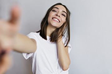 Brunette excited girl in white t-shirt making selfie. Indoor shot of emotional caucasian young lady isolated on light background.