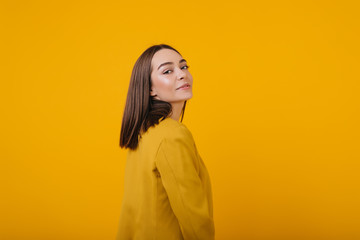 Playful girl looking over shoulder while posing on yellow background. Spectacular female model in bright jacket enjoying photoshoot.