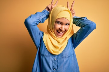 Young beautiful woman with curly hair wearing arab traditional hijab over yellow background Posing funny and crazy with fingers on head as bunny ears, smiling cheerful