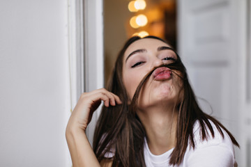 Indoor portrait of happy female model making faces during photoshoot. Photo of playful brown-haired lady posing with kissing face expression.