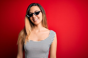 Young beautiful brunette woman wearing funny thug life sunglasses over red background with a happy and cool smile on face. Lucky person.