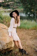 Young adorable girl in white dress and black hat, in spring, posing for camera. female portrait outdoor in mountain lake. Green fresh grass. Young unusual person enjoying nature