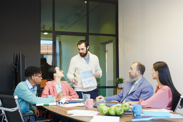 Young bearded man standing in front of his colleagues in office board room holding paper with graphs speaking on project issue