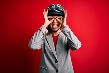 Young beautiful blonde motorcyclist woman wearing motorcycle helmet over red background doing ok gesture like binoculars sticking tongue out, eyes looking through fingers. Crazy expression.