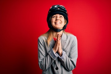 Young beautiful blonde motorcyclist woman wearing motorcycle helmet over red background begging and praying with hands together with hope expression on face very emotional and worried. Begging.