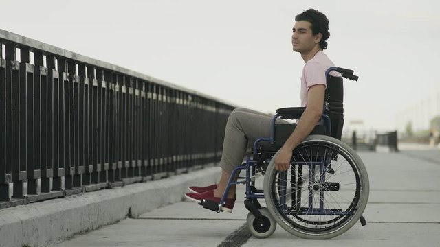 Thoughtful young man sitting on wheelchair. Sad disabled on wheelchair outside feeling lonely.