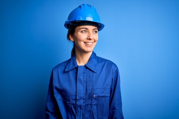 Young beautiful worker woman with blue eyes wearing security helmet and uniform looking away to side with smile on face, natural expression. Laughing confident.