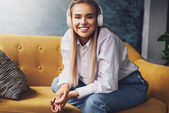 Beautiful blonde woman in white headphones sits on yellow sofa, enjoys listening to podcast. Relaxing girl in shirt