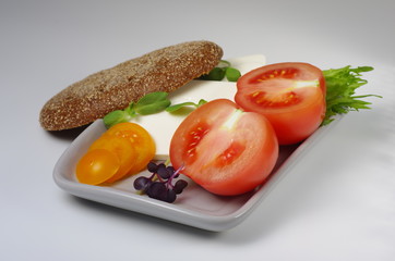 Sliced pink and yellow tomatoes and feta cheese on a plate with a piece of brown bread. Gray background. Selective focus.