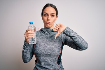 Young fitness woman wearing sport workout clothes drinking water from plastic bottle with angry face, negative sign showing dislike with thumbs down, rejection concept